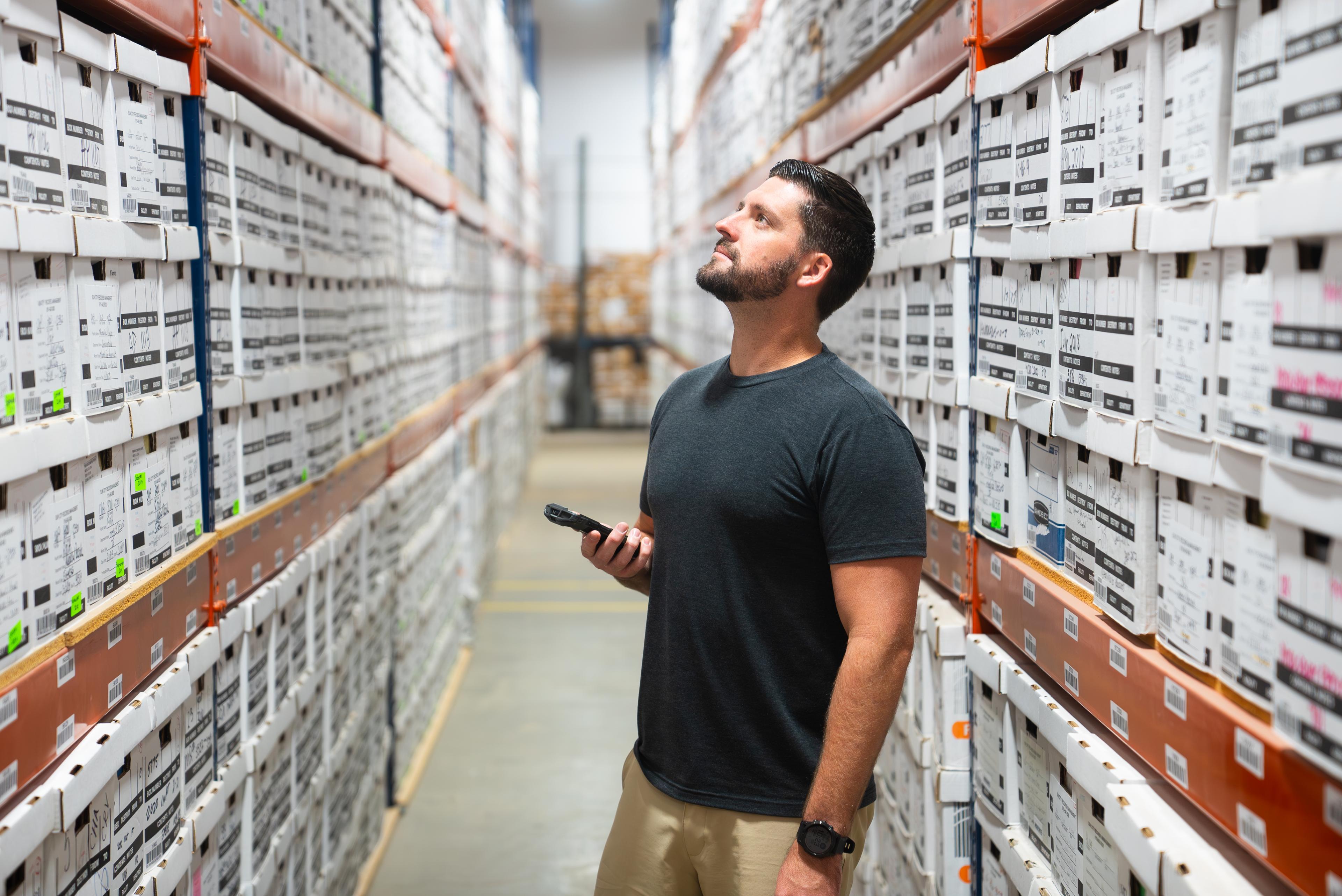 A man in an off-site physical records center holding a phone and looking at shelves of archived boxes. 